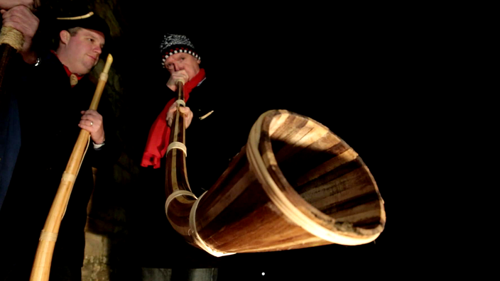 Ein Mann bläst in ein hölzernes Middwinterhorn, ein anderer steht mit einem solchen Horn daneben.