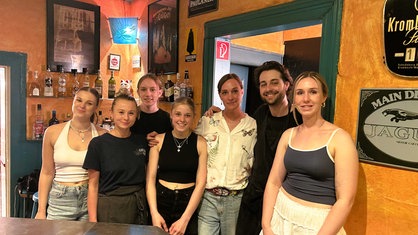 Auf dem Bild ist die Kneipenbesitzerin Julia Söhngen mit ihrem Team zu sehen. Sie stehen zu siebt hinter der Theke und lächeln in die Kamera.