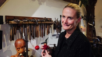 Katrin Becker hält einen kleinen gebastelten Wichtel mit langem Hut in den Händen. Sie steht in ihrer Goldschmiedewerkstatt, im Hintergrund hängen Hammer.