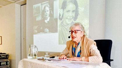 Holocaust-Überlebende Eva Weyl spricht vor einer Schulklasse. Sie trägt ein Headset. Im Hintergrund sieht man schwarz-weiß Aufnahmen von ihr als Kind. 