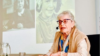 Holocaust-Überlebende Eva Weyl sitzt in einem Klassenzimmer. Sie trägt ein Headset. Im Hintergrund sieht man schwarz-weiß Aufnahmen von ihr als Kind. 