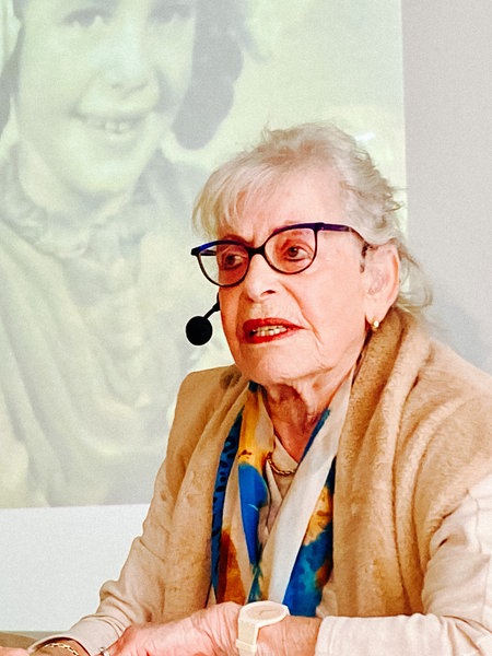 Holocaust-Überlebende Eva Weyl sitzt in einem Klassenzimmer. Sie trägt ein Headset. Im Hintergrund sieht man schwarz-weiß Aufnahmen von ihr als Kind. 