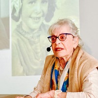 Holocaust-Überlebende Eva Weyl sitzt in einem Klassenzimmer. Sie trägt ein Headset. Im Hintergrund sieht man schwarz-weiß Aufnahmen von ihr als Kind. 