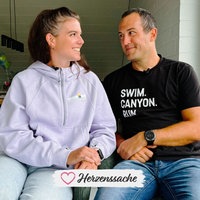 Cathrin, eine brünette Frau im lila Pullover, und Rouven, ein dunkelblonder Mann im schwarzen T-Shirt, sitzen nebeneinander und schauen sich verliebt an.