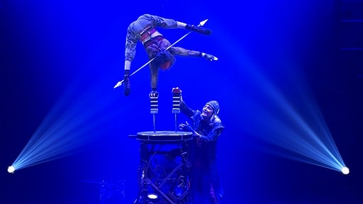 Silke Pan steht in einem inarmigen Handstand auf einer Stütze auf einem Podest in der Zirkusmanege. Unten steht ihr Mann und erhöht die zweite Stütze mit einem weiteren Baustein.