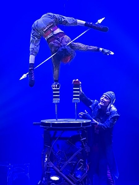 Silke Pan steht in einem inarmigen Handstand auf einer Stütze auf einem Podest in der Zirkusmanege. Unten steht ihr Mann und erhöht die zweite Stütze mit einem weiteren Baustein.