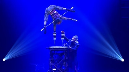 Silke Pan steht in einem inarmigen Handstand auf einer Stütze auf einem Podest in der Zirkusmanege. Unten steht ihr Mann und erhöht die zweite Stütze mit einem weiteren Baustein.