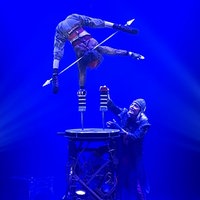 Silke Pan steht in einem inarmigen Handstand auf einer Stütze auf einem Podest in der Zirkusmanege. Unten steht ihr Mann und erhöht die zweite Stütze mit einem weiteren Baustein.