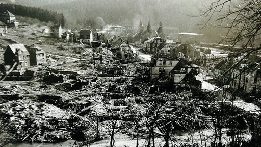 Ein schwarz-weißes Bild einer zerstörten Stadt. Häusertrümmer und Rauchschwaden sind zu sehen.