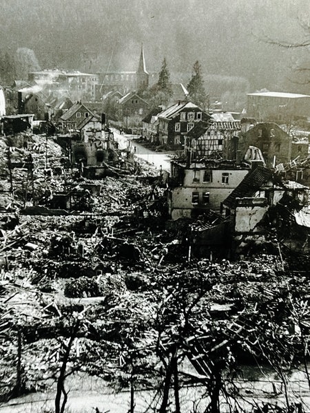Ein schwarz-weißes Bild einer zerstörten Stadt. Häusertrümmer und Rauchschwaden sind zu sehen.