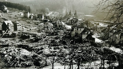 Ein schwarz-weißes Bild einer zerstörten Stadt. Häusertrümmer und Rauchschwaden sind zu sehen.