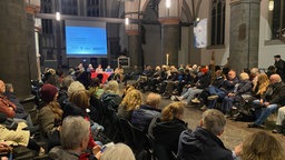 ein voll besetztes Kirchenschiff in der City Kirche in Mönchengladbach, vorne sitzten fünf Personen an einem Podium unter einer Leinwand