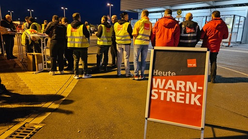 Demonstrantinnen und Demonstranten stehen vor dem Flughafen. Davor ein Schild mit der Aufschrift "verdi - Warnstreik - Heute"