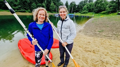 Zwei Frauen stehen an einem See. Hinter ihnen liegen bereits zwei Bords im Wasser. Die beiden halten jeweils ein Paddel in der Hand.