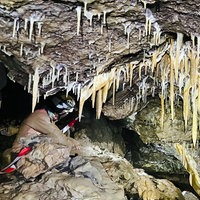 Ein Mann mit Helm und Stirnlampe in einem Durchgang in einer Höhle, von oben ragen viele Tropfsteine ins Bild