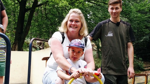Eine Mutter sitzt mit ihrem Sohn auf einem Schlitten. Sie lächelt. Hinter ihr steht Nicklas Panfilov.