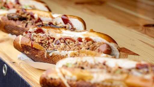 Auf einem Holzbrett liegen mehrere Hot Dogs nebeneinander, flache Weißbrote mit Wiener Wurst und Röstzwiebeln.