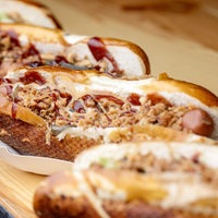 Auf einem Holzbrett liegen mehrere Hot Dogs nebeneinander, flache Weißbrote mit Wiener Wurst und Röstzwiebeln.