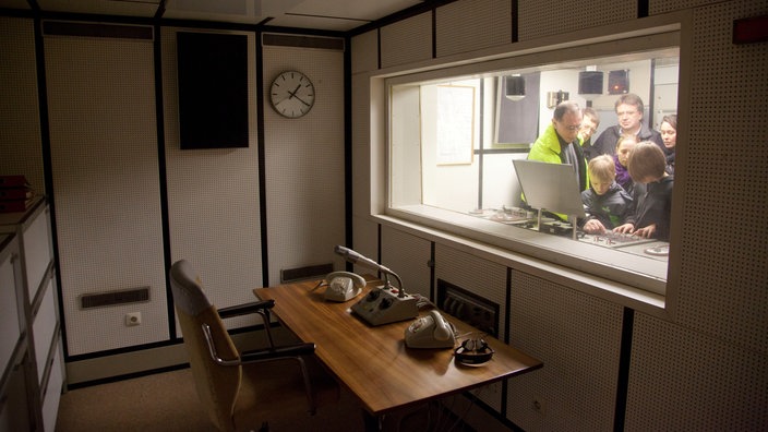 ein Raum mit einem Schreibtisch und Telefonen. Durch ein Fenster sind Menschen im Nebenraum zu sehen, die über ein Mischpult gebeugt sind