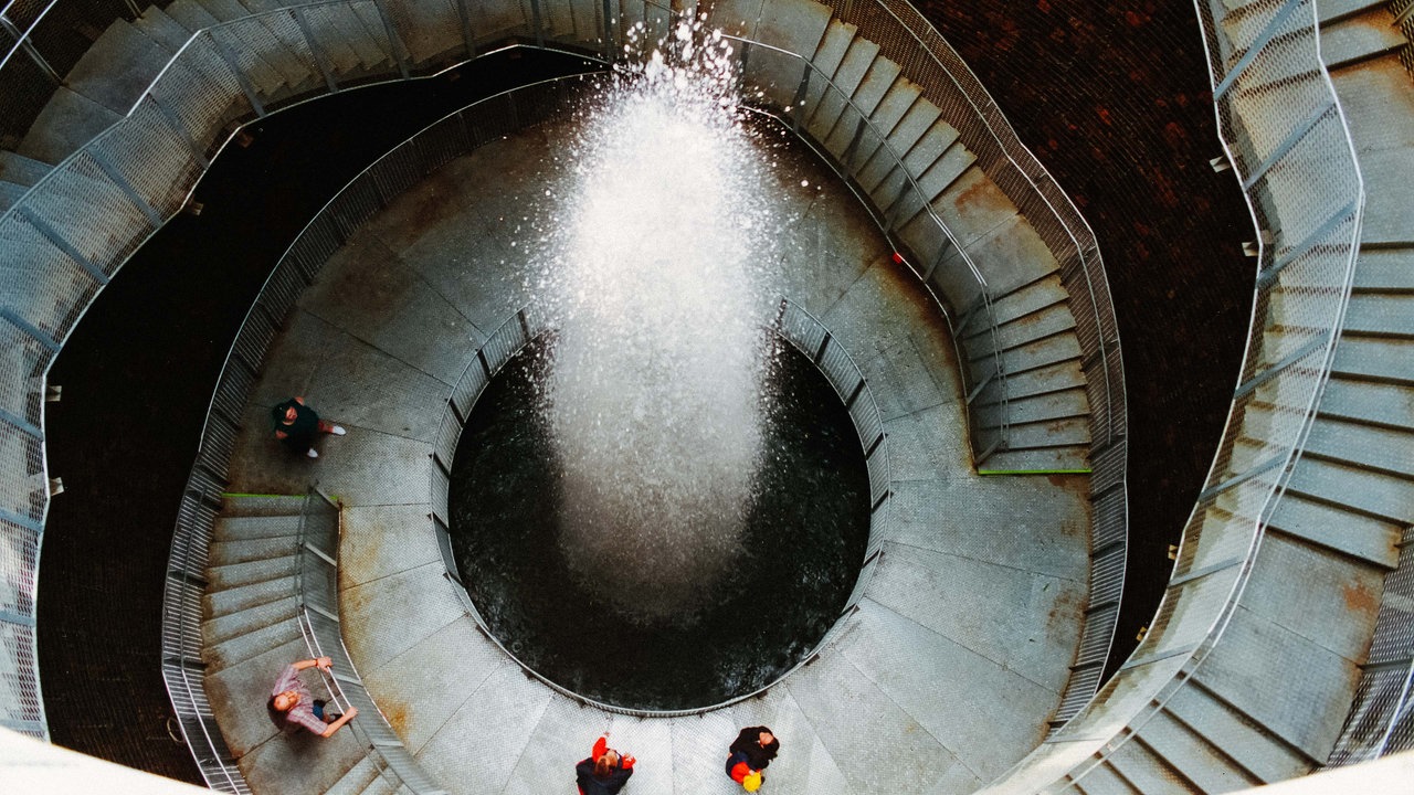 Während der Landesgartenschau Bad Oeynhausen im Jahr 2000 stehen mehrere Menschen  in einer Art Treppenhaus um einen Wasserkrater herum.