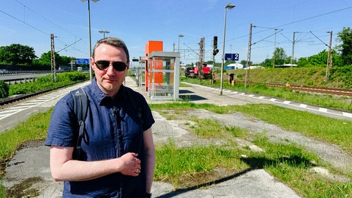 Ein Mann mit kurzen Haaren, leichtem Bart und Sonnenbrille. Er trägt ein kurzes blaues Hemd und steht an einem Bahnsteig