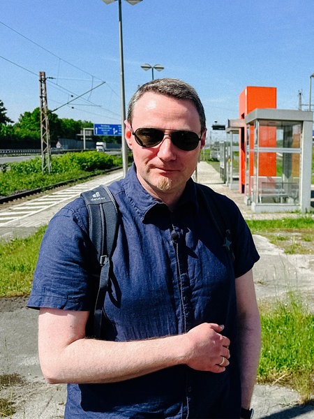 Ein Mann mit kurzen Haaren, leichtem Bart und Sonnenbrille. Er trägt ein kurzes blaues Hemd und steht an einem Bahnsteig