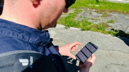 Ein Mann mit Sonnenbrille der auf sein Smartphone schaut. Im Hintergrund ein Stück Grünfläche
