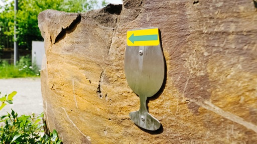 Ein brauer Stein, darauf ist eine Messingscheibe in Form eines Bierglases befestigt, auf dem Glas ist ein grüner Pfleil auf gelbem Grund zu sehenm, der nach links zeigt