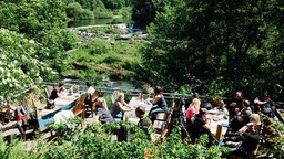Biergarten-Terrasse an einem kleinen Wasserfall. Auf der Terrasse sitzen Personen an bootsähnlichen Tischen.