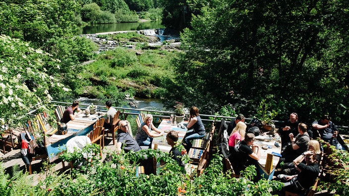 Biergarten-Terrasse an einem kleinen Wasserfall. Auf der Terrasse sitzen Personen an bootsähnlichen Tischen.