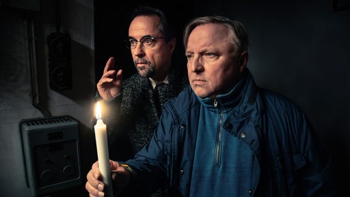 Die Schauspieler Axel Prahl und Jan Josef Liefers stehen in einem dunklen Raum, einer hält eine brennende Kerze, während beide aufmerksam in die Ferne schauen.