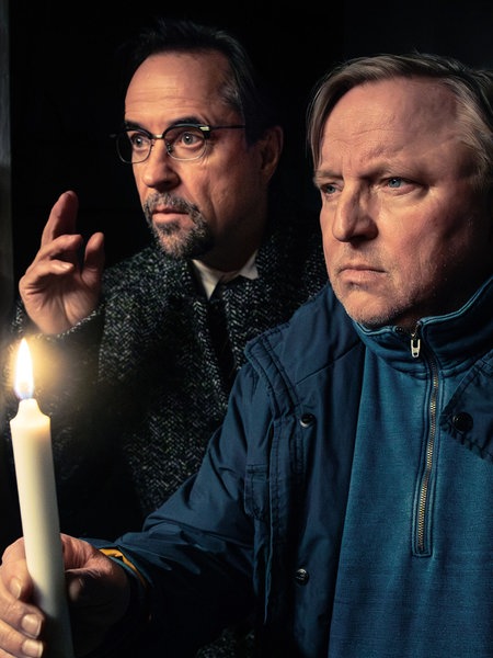 Die Schauspieler Axel Prahl und Jan Josef Liefers stehen in einem dunklen Raum, einer hält eine brennende Kerze, während beide aufmerksam in die Ferne schauen.