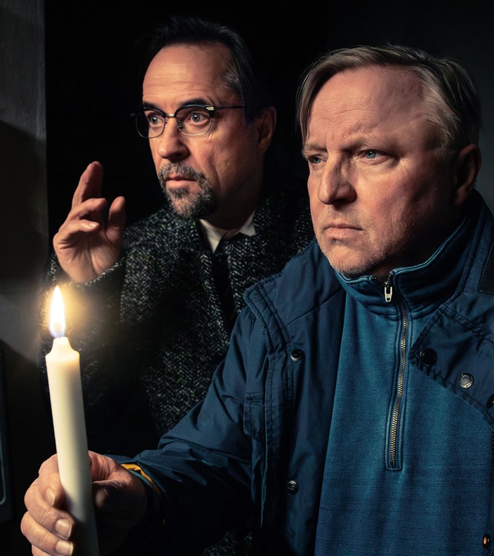 Die Schauspieler Axel Prahl und Jan Josef Liefers stehen in einem dunklen Raum, einer hält eine brennende Kerze, während beide aufmerksam in die Ferne schauen.