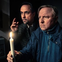Die Schauspieler Axel Prahl und Jan Josef Liefers stehen in einem dunklen Raum, einer hält eine brennende Kerze, während beide aufmerksam in die Ferne schauen.