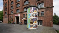 Ein großes, rotes Backsteinhaus, dessen Eingang die Überschrift "Stollwerck" trägt. Im Vordergrund steht eine bunt beklebte Litfaßsäule. 