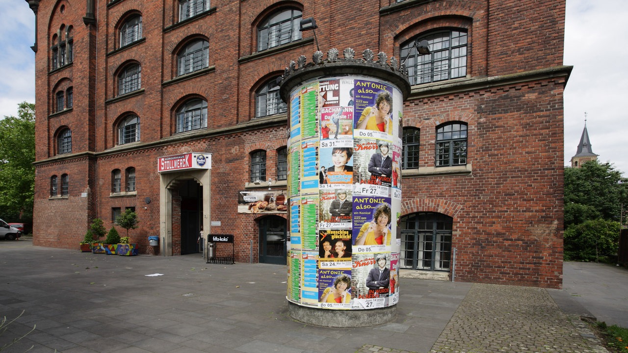 Ein großes, rotes Backsteinhaus, dessen Eingang die Überschrift "Stollwerck" trägt. Im Vordergrund steht eine bunt beklebte Litfaßsäule. 