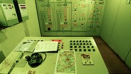 In einem Raum in einem Bunker steht eine Kontrollzentrale mit Zetteln, vielen Knöpfen und Telefonen.