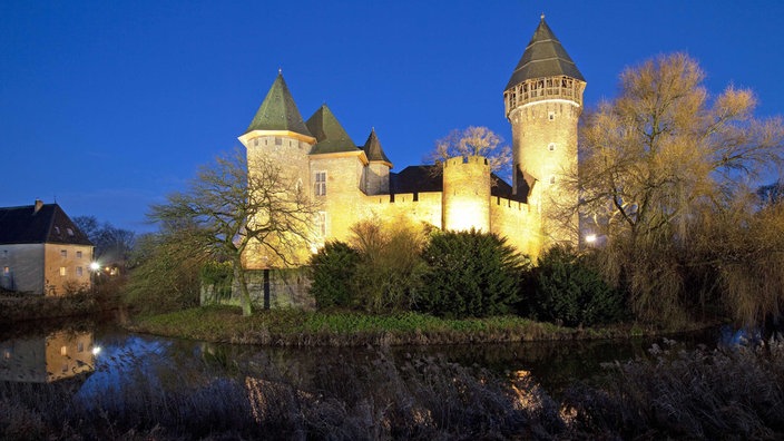 Blick auf die Burg Linn mit vier Türmen,  um die ein Wassergraben liegt, die Burg wird von gelb-weißem Licht angeleuchtet, drumherum stehen Bäume