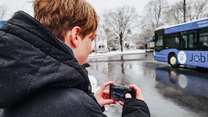 Ein Junge steht mit einem Handy in der Hand im Schnee an einem Busbahnhof 