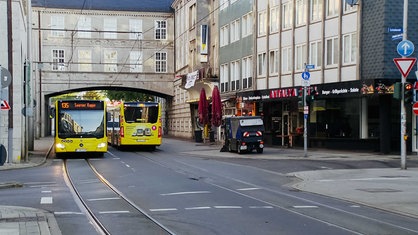 Zwei gelbe Busse der Ruhrbahn, einen sieht an von vorne, den anderen von hinten, fahren eine Straße mit Straßenbahnschienen entlang, rechts fährt ein Straßenreinigungsfahrzeug