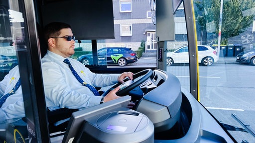 Kilian Zander, hellblaues Hemd, blaue Krawatte, schwarze Sonnenbrille, sitzt am Steuer eines Busses und blickt durch die Frontscheibe, im Hintergrund parken Autos auf der Straße