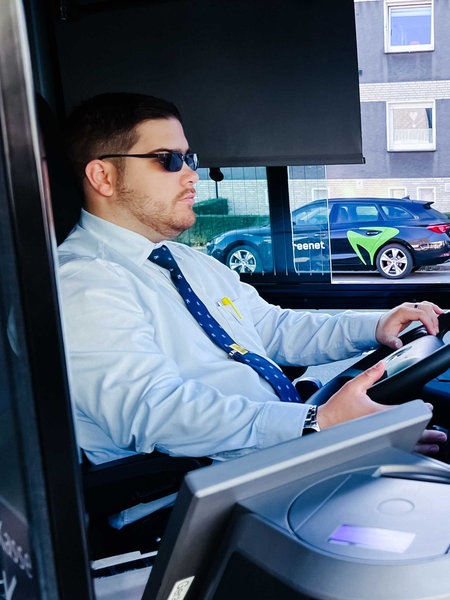 Kilian Zander, hellblaues Hemd, blaue Krawatte, schwarze Sonnenbrille, sitzt am Steuer eines Busses und blickt durch die Frontscheibe, im Hintergrund parken Autos auf der Straße