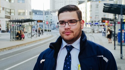 Kilian Zander, kurze braune Haare, rechteckige schwarz umrandete Brille, dunkelblaue Jacke, hellblaues Hemd, blaue Kravatte, im Hintergrund sieht man eine Bushaltestelle