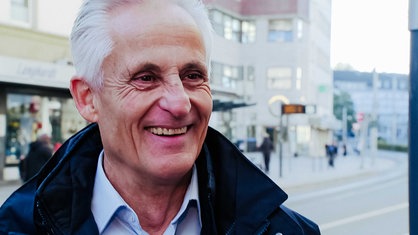 Herbert Denzelmann, kurzes, schütteres weißes Haar, breites Lächeln mit Lachfalten um die Augen, dunkelblaue Jacke, hellblaues Hemd, im Hintergrund eine Bushaltestelle