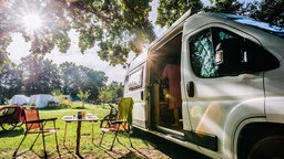 Rechts ein weißer Camping-Wagen, die Tür ist offen, links daneben stehen zwei Stühle und ein Tisch, im Hintergrund grünes Feld und Bäume