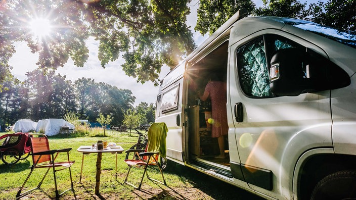 Rechts ein weißer Camping-Wagen, die Tür ist offen, links daneben stehen zwei Stühle und ein Tisch, im Hintergrund grünes Feld und Bäume