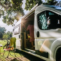 Rechts ein weißer Camping-Wagen, die Tür ist offen, links daneben stehen zwei Stühle und ein Tisch, im Hintergrund grünes Feld und Bäume