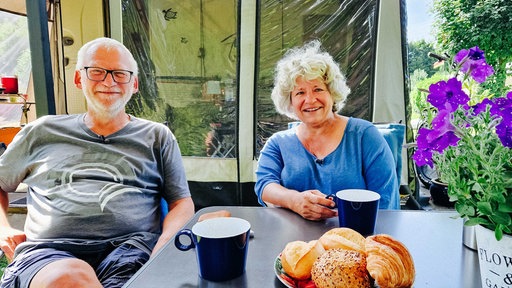 Regina Wippel und Götz Leimann sitzen nebeneinander an einem Campingtisch, darauf zwei Tassen Kaffee und ein Teller mit Backwaren.