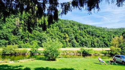 Eine Grünflache mit einem kleinen Fluss und großen, grünen Bäumen im Hintergrund. Zwei Menschen sitzen auf Campingstühlen neben einem Auto auf der Wiese.