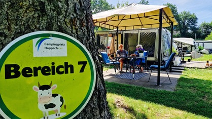 Im Vordergrung ist ein Schild an einem Baum befestigt, dass auf einen Campingbereich hinweist. Im Hintergrund erkennt man den Campingplatz von Regina Wippel un Götz Leimanns. Sie sitzen gemeinsam am Campingtisch.